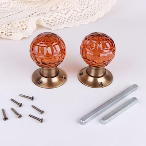 May include: Two amber-colored glass door knobs with a faceted, spherical design mounted on circular, brushed brass-toned metal bases. The set includes six small metal screws and two rectangular metal spindles for installation.