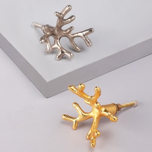 Peut inclure: Deux boutons de meuble décoratifs en forme de corail. L'un est argenté, l'autre doré. Les boutons ont une vis et une pointe blanche. Les boutons sont sur une surface gris clair.