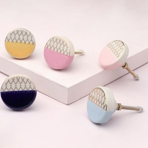 Peut inclure: Cinq boutons de placard en céramique colorés. Chaque bouton rond présente un motif géométrique doré et blanc sur la moitié supérieure. Les moitiés inférieures sont jaunes, roses, bleu marine et bleu clair. Les boutons ont des vis métalliques.