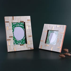 Op de afbeelding: Twee decoratieve fotolijsten. De lijsten hebben een rechthoekige vorm met een mozaïkpatroon in crème en goud. De lijsten hebben een groen-witte achtergrond. Ze staan op een donkergroen oppervlak.