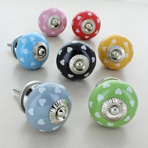 Könnte beinhalten: Satz von zehn Keramik-Schrankknöpfen mit weißen Herzmustern. Die Knöpfe sind in verschiedenen Farben, darunter rosa, rot, schwarz, gelb, blau und grün.