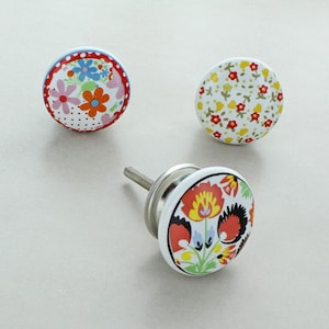 May include: Three ceramic cabinet knobs with floral patterns. One has a red and white polka dot border with colourful flowers. Another has a yellow background with red and yellow flowers. The third knob has a vibrant floral design.