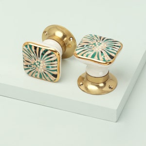 Peut inclure: Deux boutons de meuble carrés avec une base blanche, une bordure dorée et un motif floral vert, noir et beige. Les boutons ont une base en laiton avec des trous de vis, sur fond blanc.