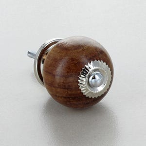 May include: A wooden cabinet knob with a round, brown finish. The knob features a silver-coloured, decorative rosette and a threaded screw for installation. Suitable for drawers and cabinets.