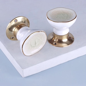 May include: Two white ceramic door knobs with gold trim and brass bases. The knobs have a crackled, aged appearance on the white surface. The brass bases are round with screw holes for mounting. The knobs are designed for cabinet or drawer use.