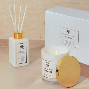 Könnte beinhalten: Ein weißes Duftgeschenkset mit einem Schilf-Diffusor, einer Duftkerze und einer Box. Der Schilf-Diffusor und die Kerze haben goldene Akzente und Etiketten mit der Aufschrift "New Book". Die Kerze ist angezündet, und die Box trägt die Aufschrift "Fragrance Gift Set".