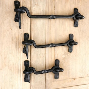 May include: Three black, wrought iron door latches are mounted on a light wood surface. Each latch features a curved arm with a decorative knot and a hook. The latches are designed for securing doors or gates.