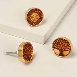 May include: Three decorative cabinet knobs with brass-colored rims and wooden faces. The wooden faces feature intricate designs, including a tree, a circular pattern, and a leaf-like pattern. The knobs are designed for easy installation.