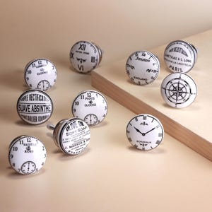 Op de afbeelding: Een verzameling wit en zwart decoratieve kastknoppen met klokgezichten en diverse teksten. De knoppen hebben ontwerpen zoals kompassen, klokgezichten en teksten zoals "London" en "Paris".