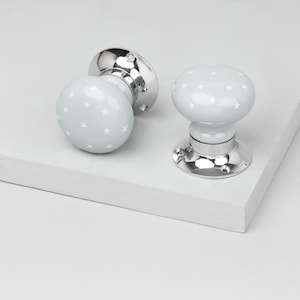 May include: Two door knobs with a light gray finish and white star pattern. The knobs have a rounded shape and are mounted on a shiny silver base. The door knobs are on a white surface.