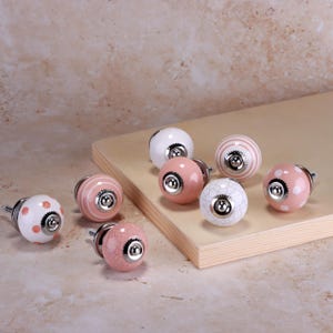 Peut inclure: Ensemble de boutons de meubles décoratifs en céramique de différents motifs. Les boutons présentent un mélange de couleurs, dont le blanc, le rose et le crème, avec des motifs tels que des rayures, des pois et des craquelures. Chaque bouton a une base en métal argenté.