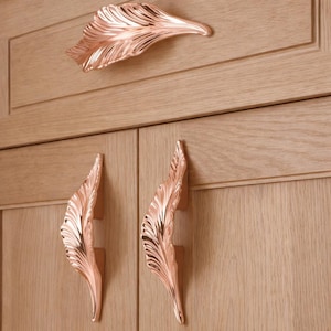 Könnte beinhalten: Schrankbeschläge in Roségold mit Blattdesign. Das Bild zeigt einen einzelnen Griff über zwei passenden Griffen. Die Griffe sind an einem hellbraunen Holzschrank befestigt. Das Blattdesign hat detaillierte Adern.
