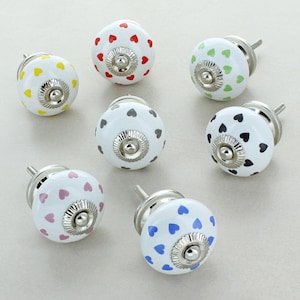 May include: Set of 7 ceramic cabinet knobs with a white base and different coloured hearts. The knobs have a silver metal screw and a silver metal ring around the base.