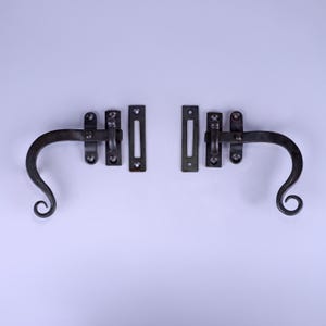 May include: Two wrought iron latches with a dark, aged finish. Each latch has a curved handle with a scroll design, a pivoting arm, and a rectangular keeper. The hardware is designed for a rustic or vintage aesthetic.