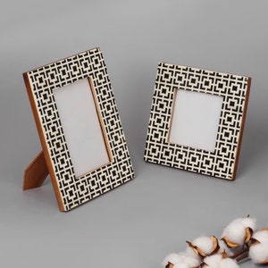 Może przedstawiać: Dwa geometryczne ramki do zdjęć w kolorze czarno-białym. Większa ramka stoi pionowo, a mniejsza leży płasko. Obie ramki mają białe tło z czarnym geometrycznym wzorem.