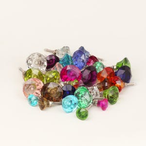 May include: A collection of colorful, faceted glass cabinet knobs in various jewel tones, including pink, blue, green, and purple. Each knob has a silver metal screw for easy installation. The knobs are arranged in a pile.