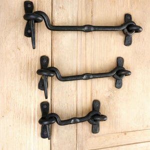 Op de afbeelding: Drie zwarte smeedijzeren poortgrendels. Elke grendel heeft een gebogen arm met een decoratieve knoop en een haak. De grendels zijn gemonteerd op een lichtgekleurd houten oppervlak.