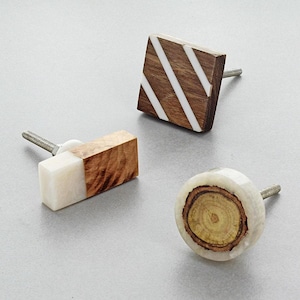 Op de afbeelding: Drie decoratieve kastknoppen. Eén is vierkant met diagonale witte strepen op een bruine houten basis. Een andere is rechthoekig, half wit en half bruin hout. De derde is rond met een houtnerfcentrum.