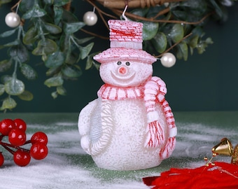 Pode incluir: Uma vela em forma de boneco de neve com cachecol, chapéu e acabamentos vermelhos e brancos. A vela tem um rosto sorridente com um nariz vermelho e é rodeada por elementos decorativos, incluindo uma grinalda e bagas. Uma decoração festiva.