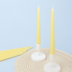 Può includere: Due candele affusolate gialle in portacandele bianchi sono esposte su un tappetino intrecciato beige su sfondo azzurro. Diverse altre candele gialle sono disposte a sinistra. Le candele hanno un design classico ed elegante.