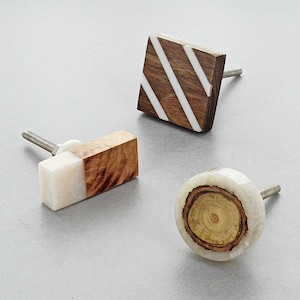 May include: Three wooden cabinet knobs with different designs. The first knob is square with white stripes on a brown background. The second knob is rectangular with a white top and brown bottom. The third knob is round with a brown centre and a white outer ring.