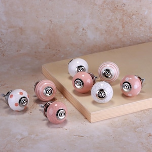 Könnte beinhalten: Eine Sammlung von Keramik-Schrankknöpfen in verschiedenen Designs und Farben. Die Knöpfe weisen Muster wie Punkte, Streifen und Craquelé-Oberflächen auf. Die Farbpalette umfasst Weiß, Rosa und silberfarbene Hardware. Diese dekorativen Knöpfe sind für Möbel und Heimdekoration konzipiert.