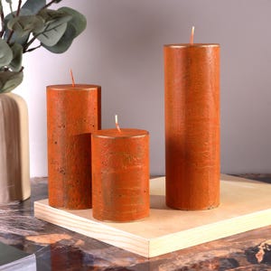 Peut inclure: Trois bougies piliers orange de différentes hauteurs. Les bougies ont une surface texturée et un bord doré métallique. Chaque bougie a une mèche blanche. Les bougies sont exposées sur une surface en bois de couleur claire.