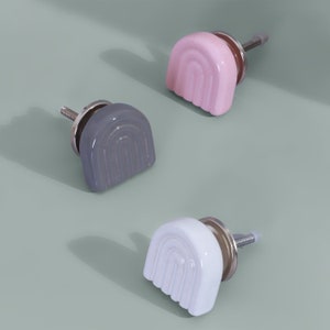 Puede incluir: Tres pomos de armario de cerámica con forma de arcoíris. Los pomos son de color rosa, gris y blanco.