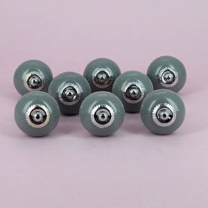 May include: Eight grey ceramic cabinet knobs with a crackle finish. Each knob has a silver-coloured metal base and decorative silver trim. The knobs are arranged on a light pink background, suitable for drawers or cupboards.