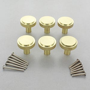 May include: Six brass cabinet knobs with a circular top and a textured base. The knobs are arranged in two rows of three, with a set of screws on each side. These hardware pieces are ideal for updating furniture.