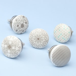May include: Five decorative cabinet knobs with various patterns. The knobs are white with grey designs, including floral, geometric, and striped patterns. Each knob has a silver metal base and stem, set against a light blue background.