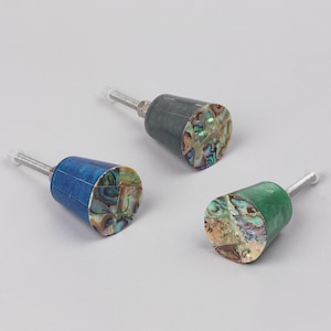 May include: Three cabinet knobs with a cylindrical design. One is blue, one is green, and one is grey. Each knob features an iridescent abalone shell inlay on the face. They have a threaded metal stem.