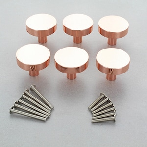 Puede incluir: Seis pomos de armario de oro rosa con un diseño plano y circular. Cada pomo tiene un vástago cilíndrico corto y va acompañado de un juego de tornillos plateados. Los pomos están dispuestos en dos filas de tres sobre una superficie gris claro.