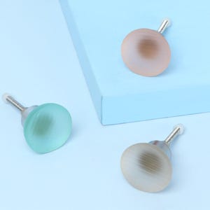 Può includere: Tre pomelli decorativi per mobili con un design rotondo e opalescente nei toni del pesca, verde e beige. Ogni pomello ha uno stelo color argento e una punta bianca. I pomelli sono disposti su una superficie azzurra.