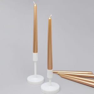 Puede incluir: Dos velas doradas cónicas en soportes blancos están de pie, con varias más tumbadas. Las velas tienen un acabado metálico y están sobre un fondo gris claro. Los soportes tienen un diseño minimalista.