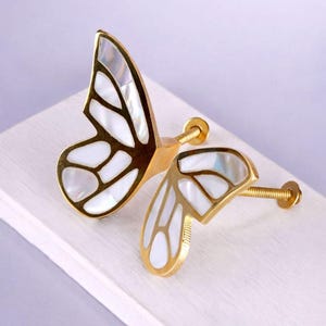 May include: Butterfly-shaped cabinet knobs with gold-colored metal frames and white, iridescent inlays. The knobs have a screw for easy installation. These decorative knobs add a unique touch to furniture and are ideal for a dresser or cabinet.