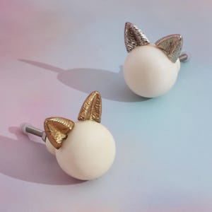 Può includere: Due pomelli decorativi per mobili con base sferica bianca e accenti metallici dorati e argentati a forma di orecchie di coniglio. Su uno sfondo sfumato pastello rosa e blu.
