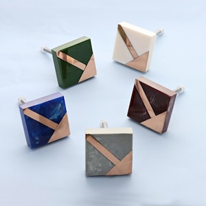 Peut inclure: Cinq boutons de placard carrés de différentes couleurs : vert, blanc, rouge foncé, bleu et gris. Chaque bouton présente un motif géométrique de couleur cuivre. Les boutons sont montés sur des vis argentées, adaptés aux meubles ou à la décoration.
