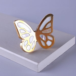 May include: A gold-colored butterfly-shaped decorative object with white iridescent accents. The butterfly is resting on a white surface, with a soft purple background.