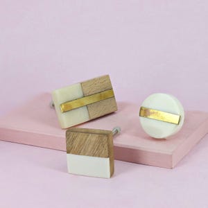 Pode incluir: Três puxadores decorativos para armários em várias formas. Um é retangular com secções em madeira e creme e um detalhe dourado. Outro é um puxador redondo com um detalhe dourado. O terceiro é um puxador quadrado com secções em madeira e creme.