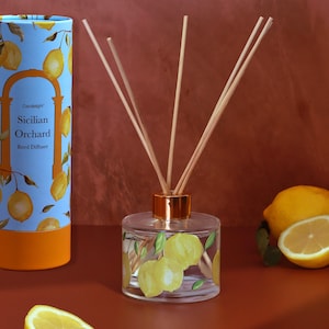 Puede incluir: Un difusor de caña de vidrio transparente con un collar dorado y cañas de madera. El difusor está decorado con limones amarillos y hojas verdes. Una caja cilíndrica con las palabras "Candlelight Sicilian Orchard Reed Diffuser" está en el fondo.