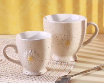 May include: Two beige ceramic mugs with a daisy design. The mugs have a speckled finish and a white interior. One mug is larger than the other. A silver spoon rests on the table next to the mugs. The background is a yellow wall.