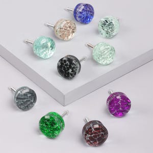 Può includere: Una collezione di pomelli per mobili in vetro colorato con effetto craquelé. I pomelli sono in vari colori, tra cui blu, verde, nero e viola, e hanno steli argentati. I pomelli sono disposti su una superficie grigio chiaro.