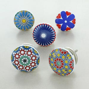 May include: Five decorative ceramic cabinet knobs with colorful geometric patterns. The knobs feature designs in blue, red, yellow, and green, with silver hardware. The knobs are round and have a variety of patterns.