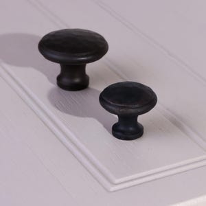 May include: Two round cabinet knobs with a matte black finish and a hammered, textured surface. Each knob features a circular top and a tapered base, designed for attachment to furniture or cabinetry doors and drawers.