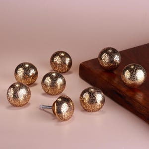 Può includere: Otto pomelli dorati per mobili con intricati motivi floreali. Un pomello è attaccato a una vite argentata. I pomelli sono disposti su una superficie rosa chiaro e una tavola di legno.