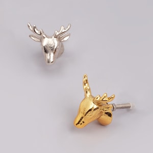 Pode incluir: Dois puxadores decorativos para armários em forma de cabeça de veado. Um é prateado e o outro dourado. Ambos os puxadores são provavelmente de metal e projetados para serem fixados a um armário ou gaveta. Medidas em centímetros.