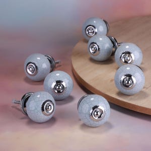 Peut inclure: Ensemble de huit boutons de meuble en céramique bleu clair avec un fini craquelé. Chaque bouton est doté d'une base et d'une quincaillerie en métal argenté. Présentés sur une surface en bois.