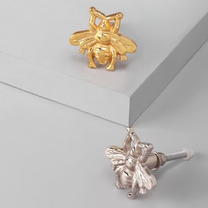 Könnte beinhalten: Zwei möbelförmige Knöpfe in Bienenform, einer goldfarben und einer silberfarben. Die Knöpfe sind aus Metall gefertigt und weisen detaillierte Flügel und Körper auf. Der goldfarbene Knopf befindet sich über dem silberfarbenen Knopf.