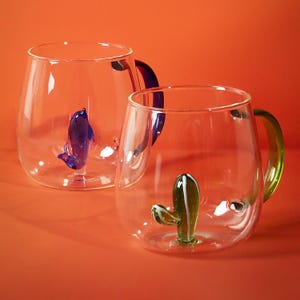 Può includere: Due tazze di vetro trasparente con corpi arrotondati e manici colorati. Una tazza ha un delfino blu all'interno, l'altra un cactus verde. I manici sono viola e verdi. Le tazze poggiano su una superficie arancione.